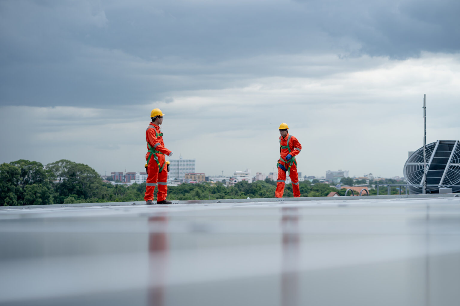 Two men in orange safety gear stand on a roof. The sky is cloudy and the city below is visible